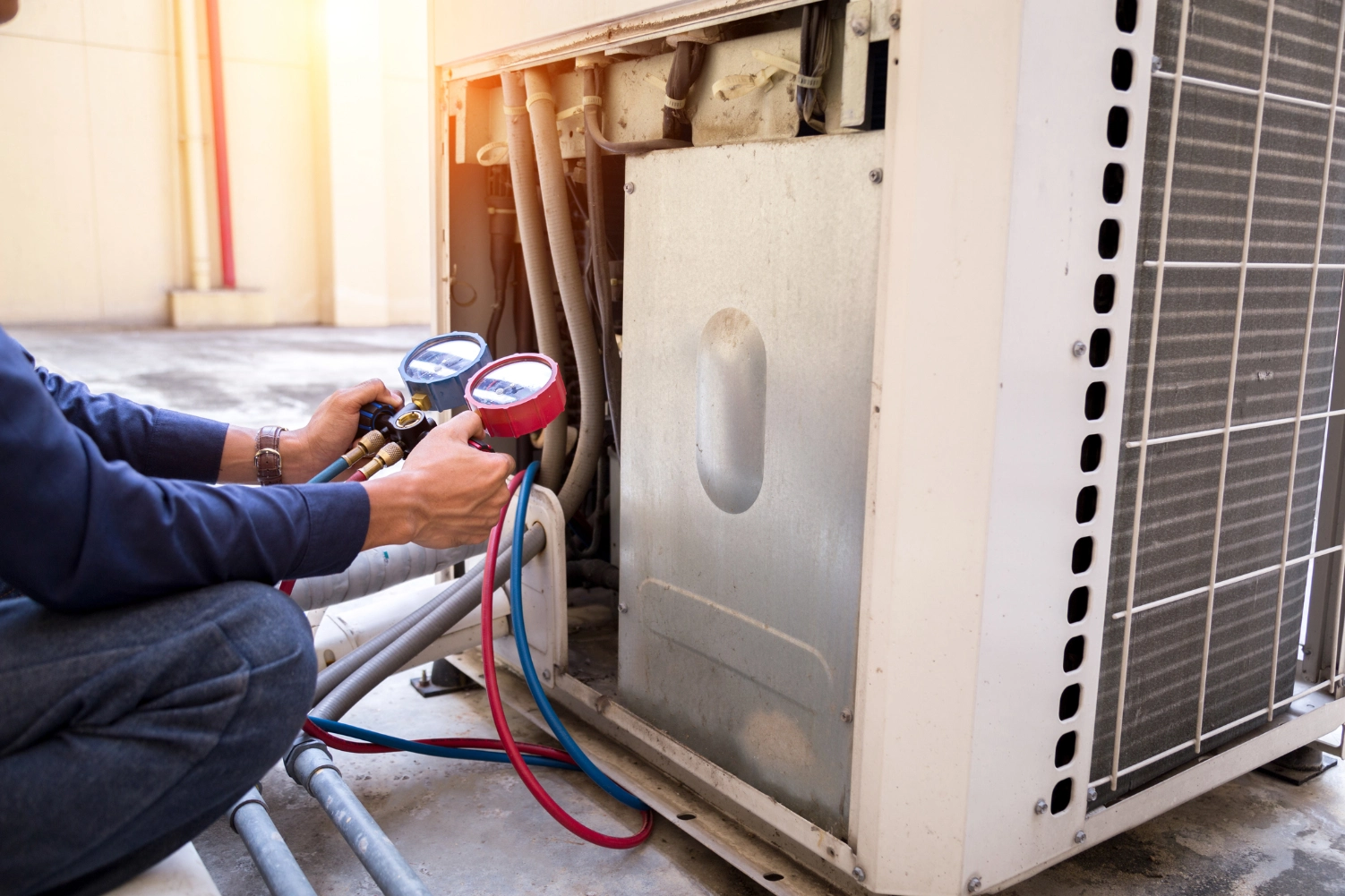 Man working on an air conditioner with a pressure gauge, surrounded by pipes and a wall in the background