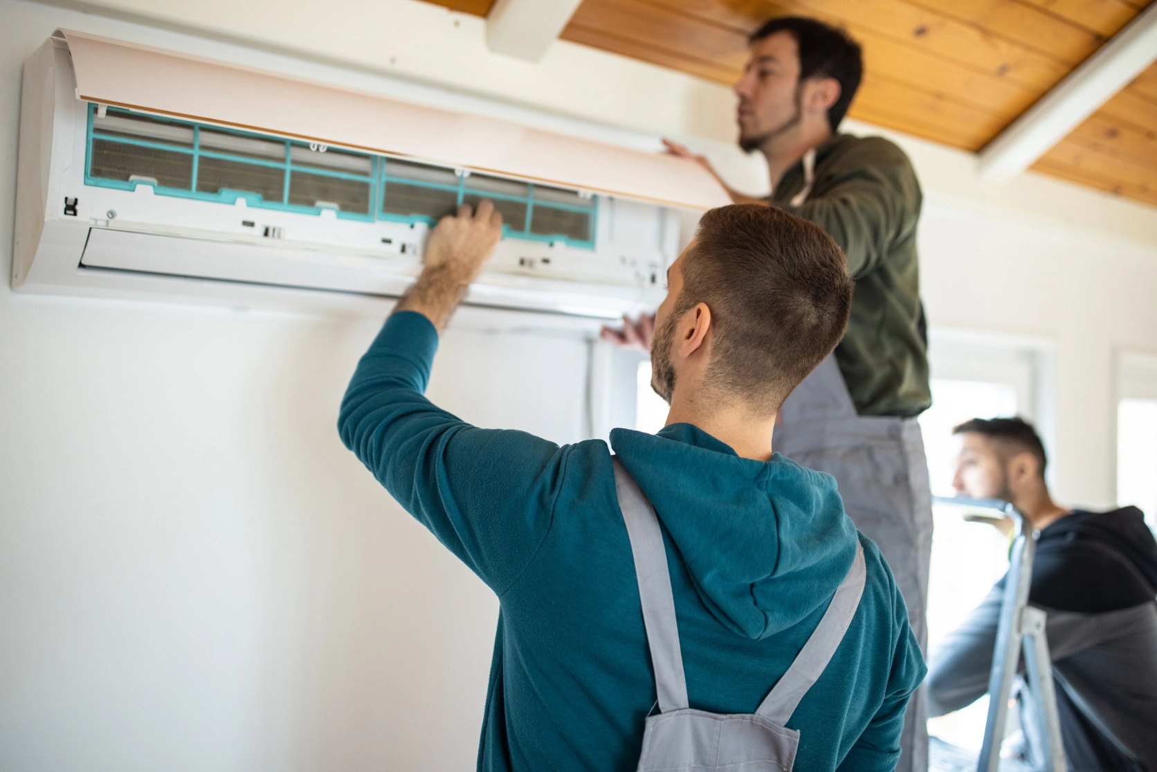 Two men working on an air conditioner in a room, with one of them standing on a ladder