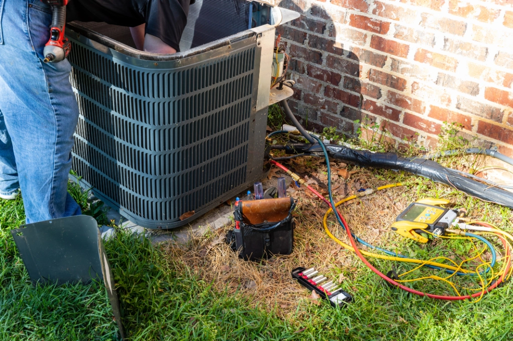 Man working on an air conditioner outside of a house, surrounded by tools and wires on the grass