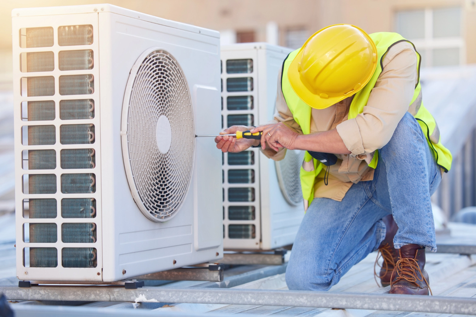 Man in a hard hat and safety vest working on an air conditioner, holding a tool in his hand