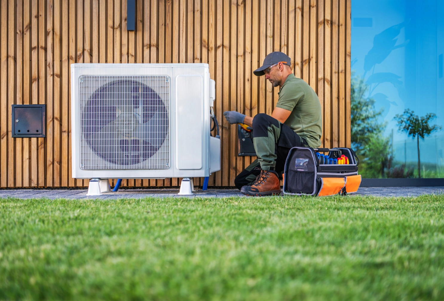 Man wearing a cap and gloves, sitting on the grass in front of an air conditioner