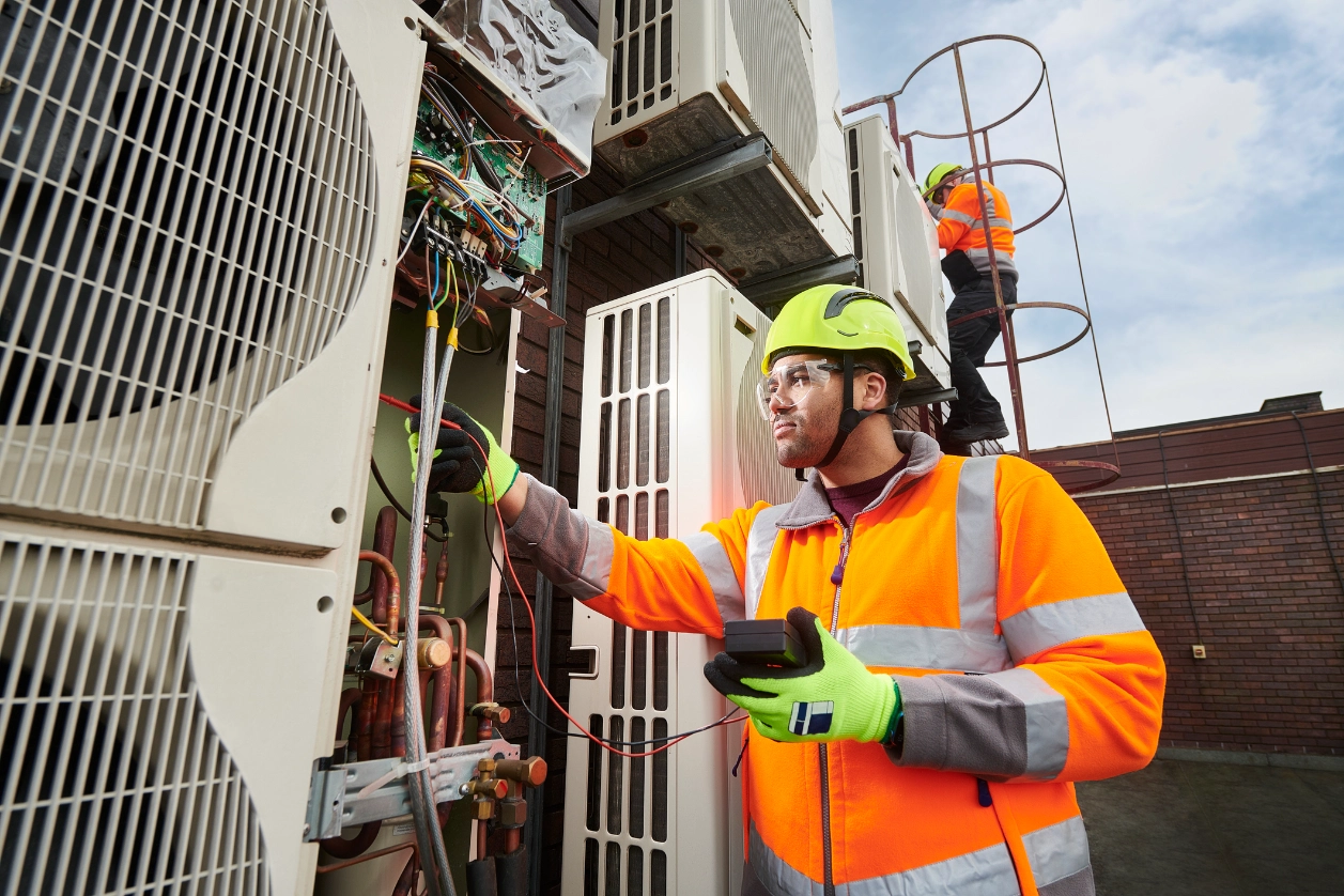 Man in an jacket and green helmet working on an air conditioner, holding a device in his hand