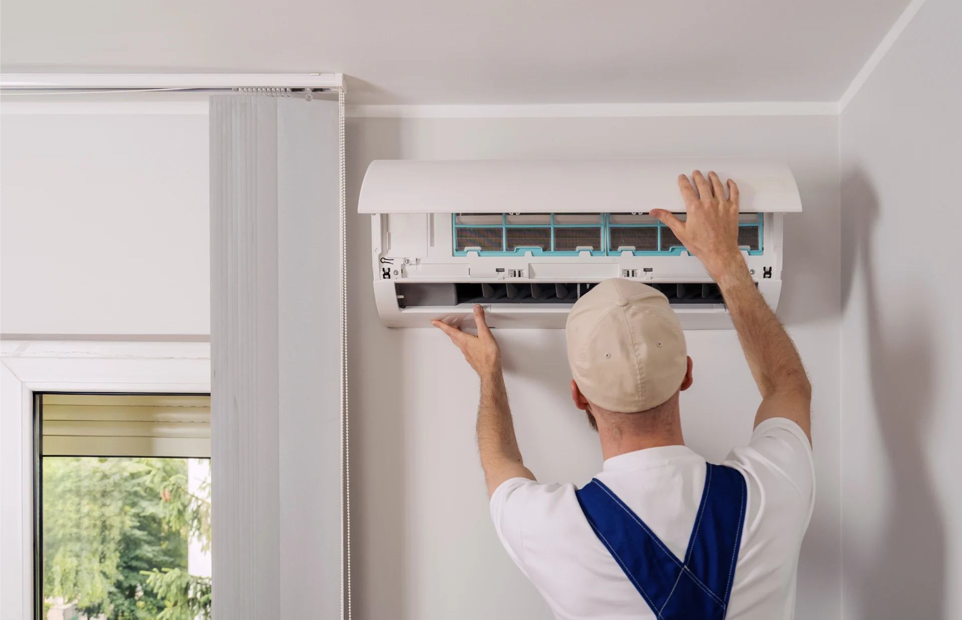 Man installing an air conditioner on the wall, with a window to the left and trees visible outside