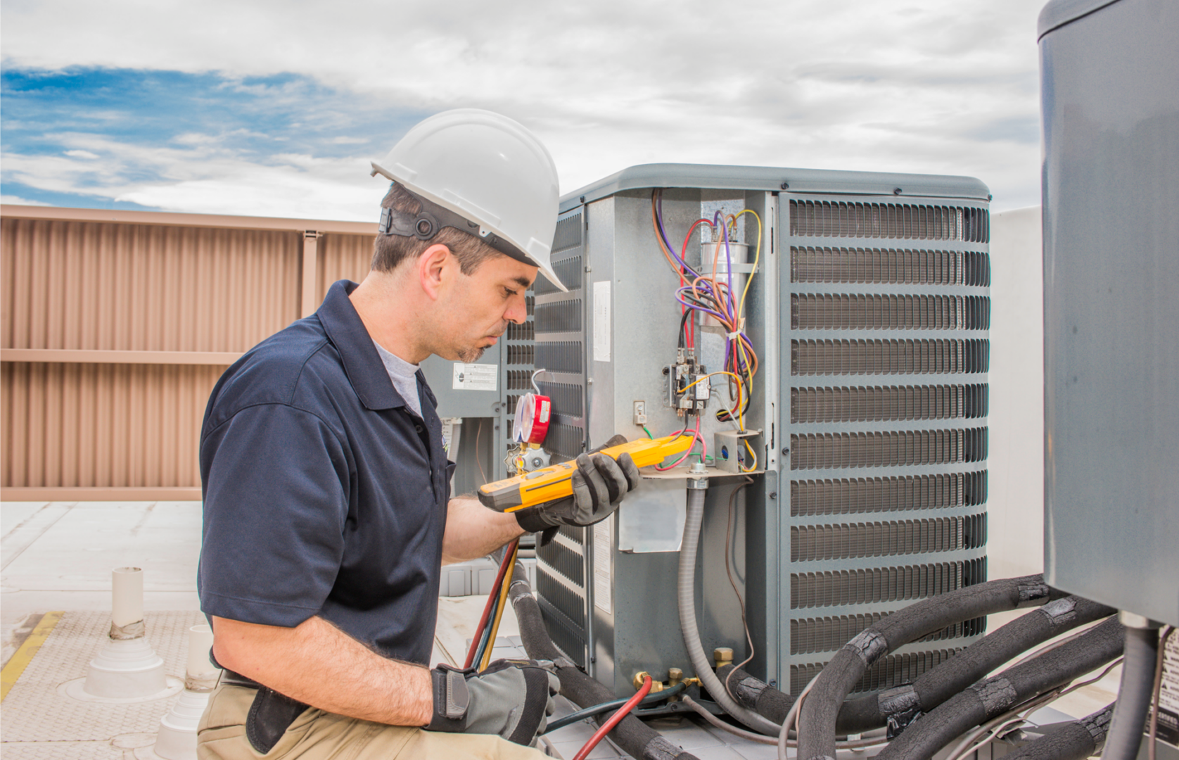 Man in a hard hat working on an air conditioner, holding a tool in his hand