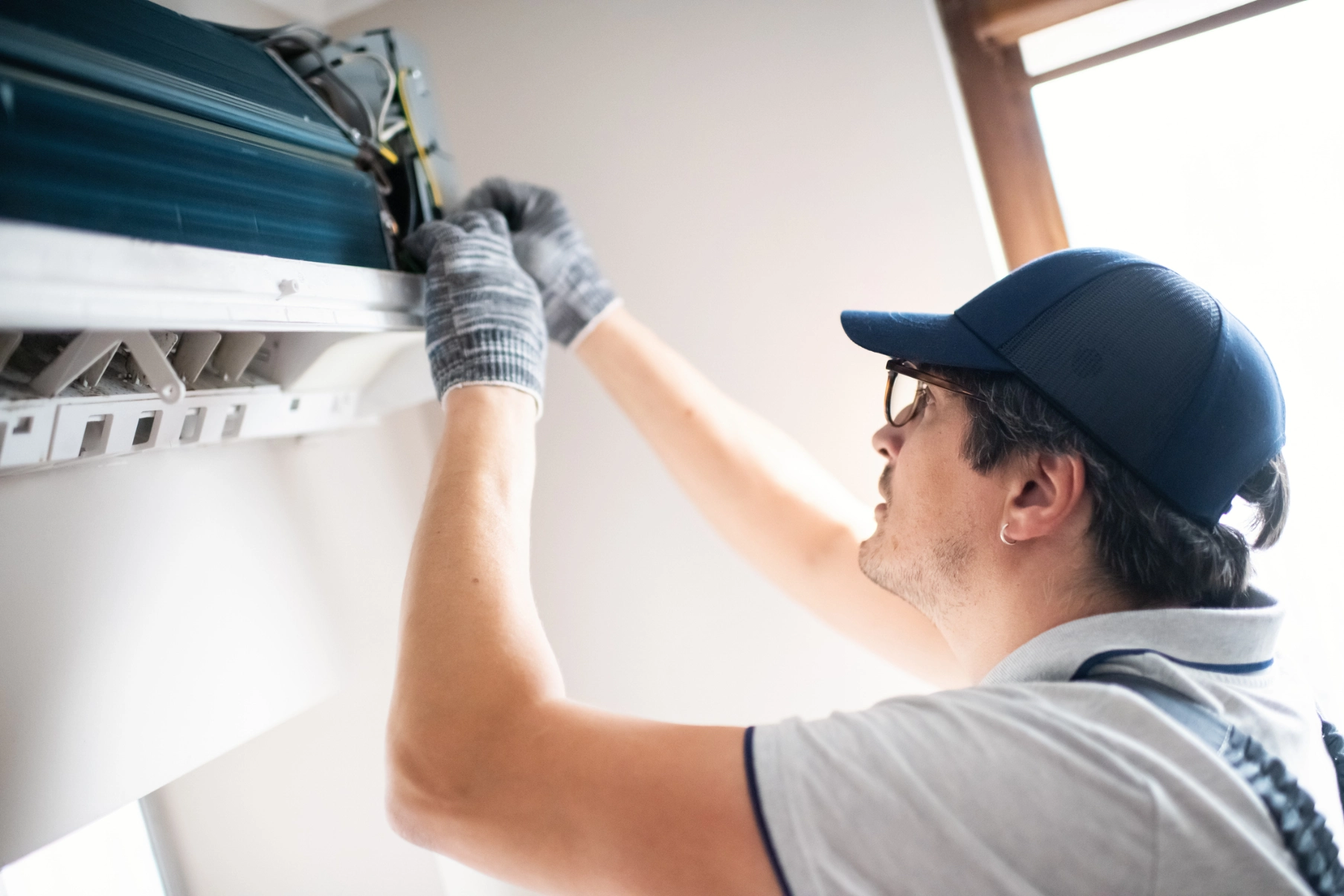 Man wearing a cap and gloves, working on an air conditioner in front of a wall with a window